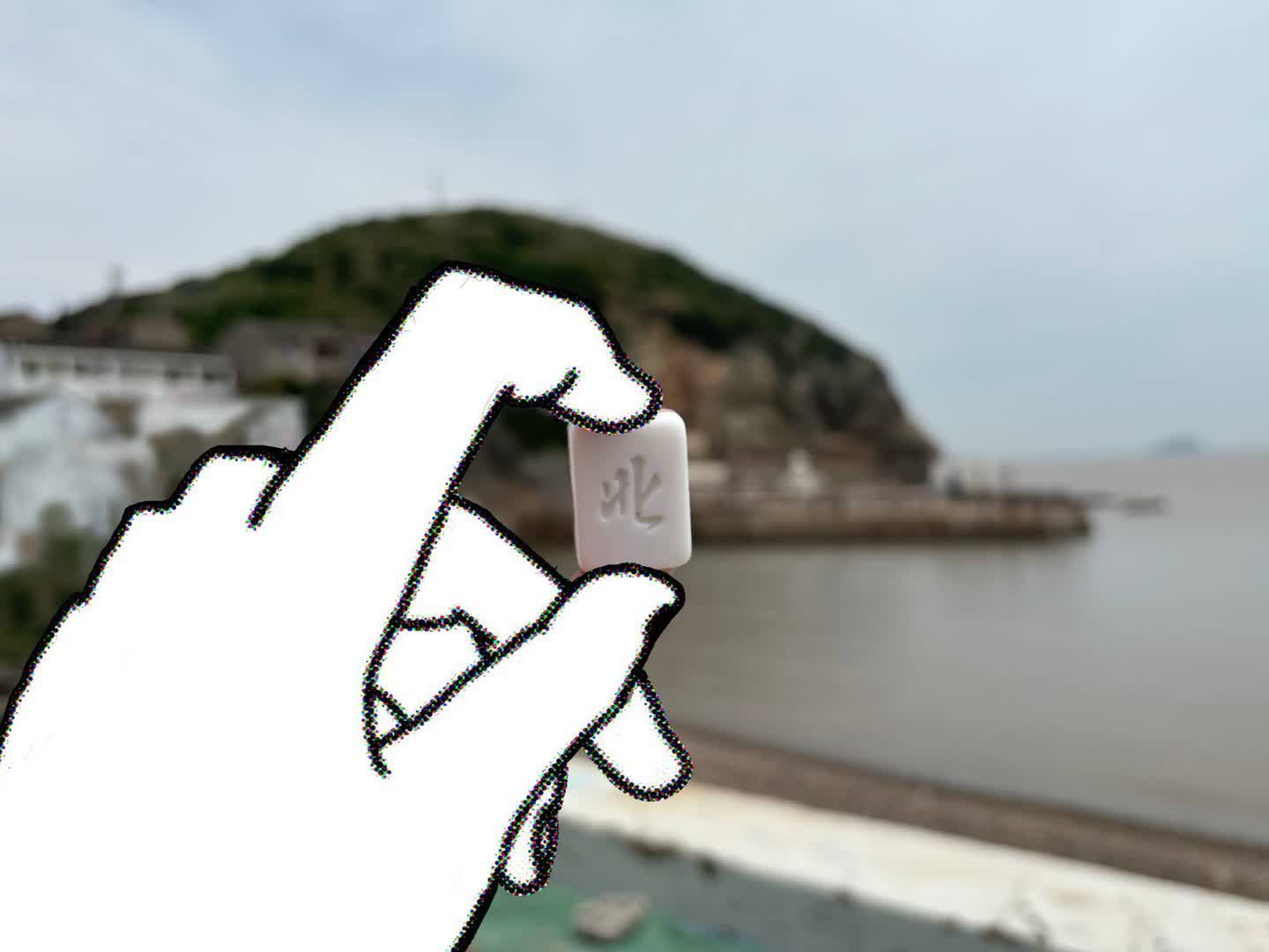 手部特寫,舉著一個麻將牌,寫著北;背景是島、石灘和海