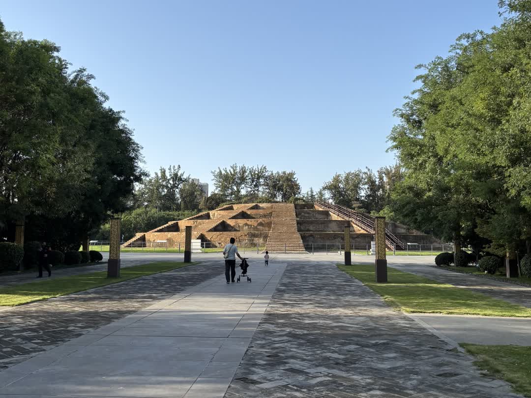 畫面以石板路為引導線，指向遠處棕色的天壇。背景是晴朗的天空和茂密的綠色樹木。有成人和兒童在石板路上散步。